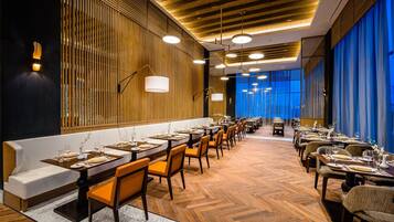 A restaurant with long wooden tables and orange chairs, a marble floor, and a blue curtain backdrop.