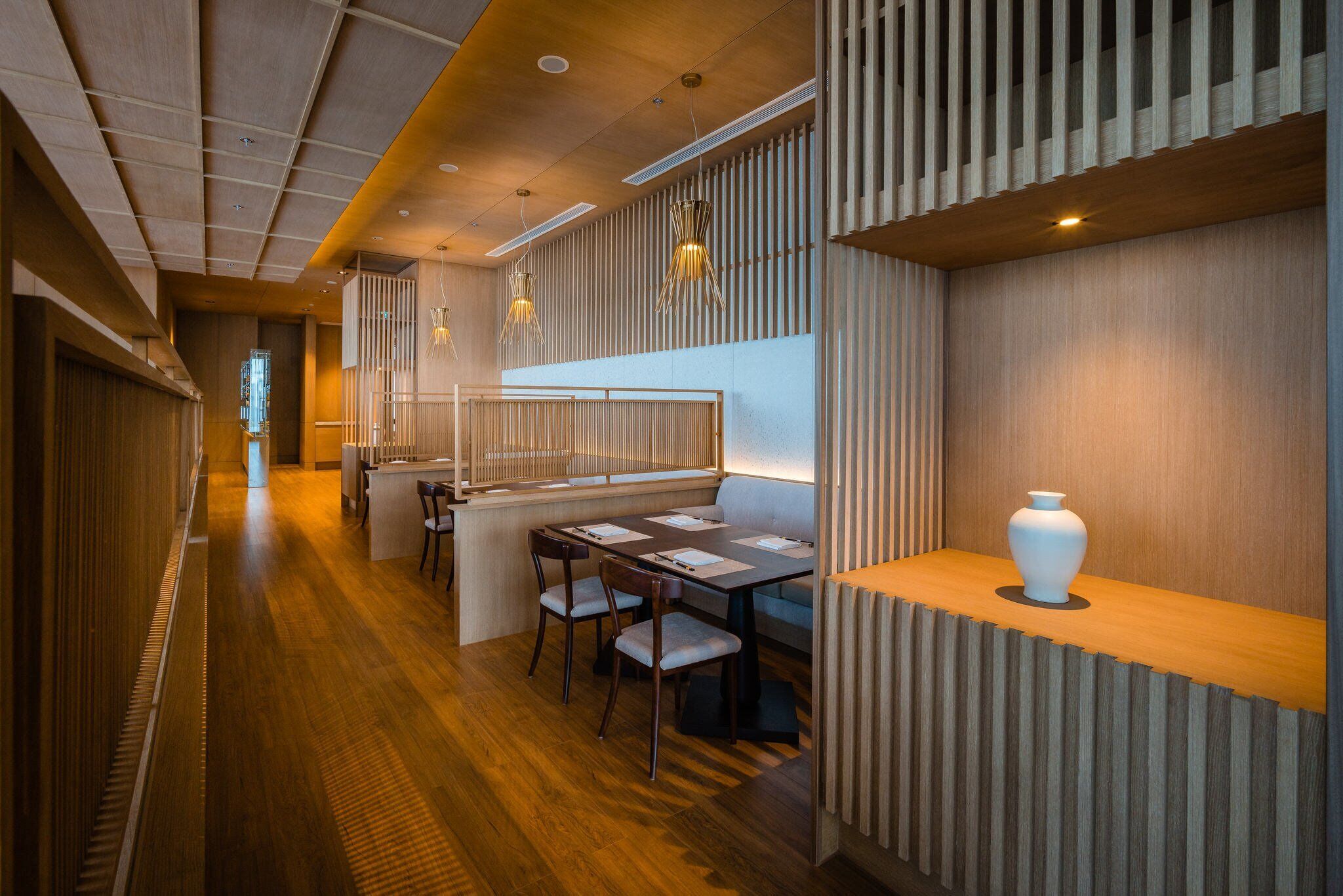Un interior moderno con suelo de madera, un área de estar con mesas y sillas, y un jarrón decorativo sobre un repisa de madera.