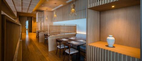 Un interior moderno con suelo de madera, un área de estar con mesas y sillas, y un jarrón decorativo sobre un repisa de madera.