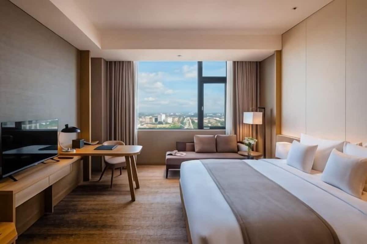 Ein Hotelzimmer mit Bett, Schreibtisch, Stuhl, Fernseher und einem Fenster mit Blick auf die Stadt.