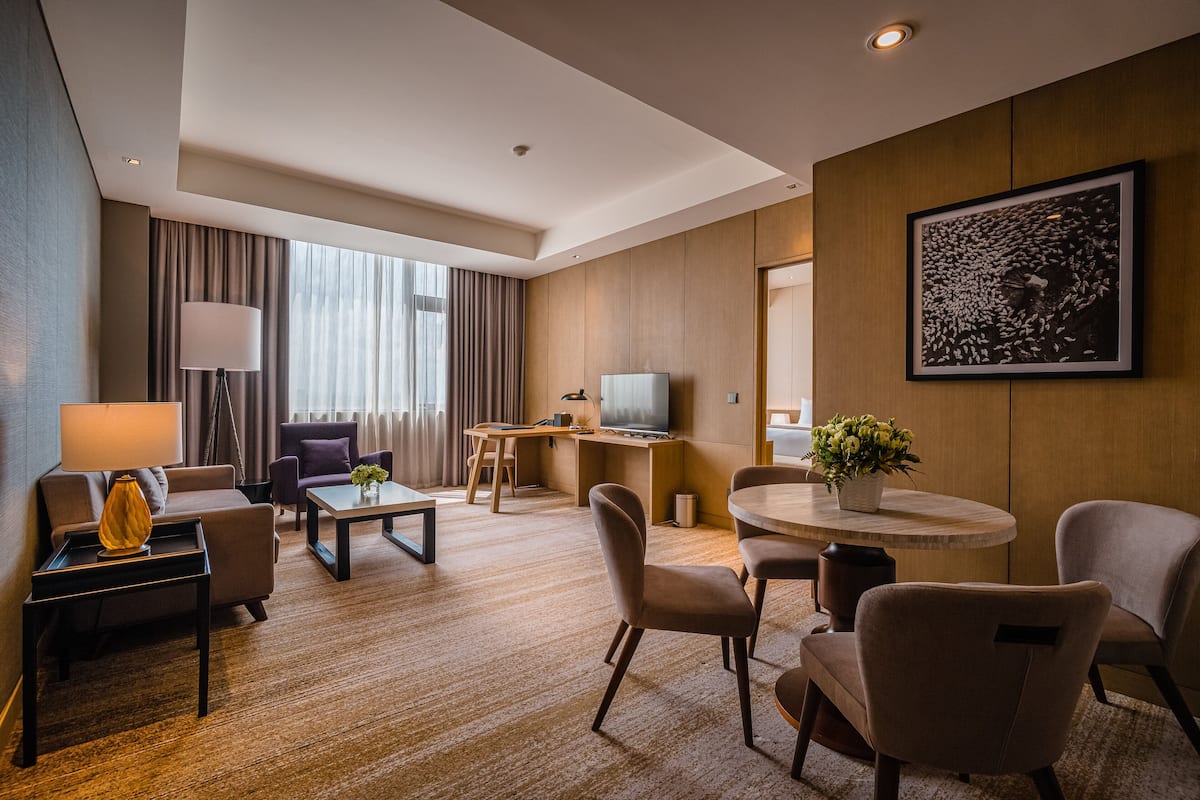 Ein modernes Hotelzimmer mit einer Sitzecke, Sesseln, einem Esstisch, einem Fernseher und einem großen Fenster mit Vorhängen.