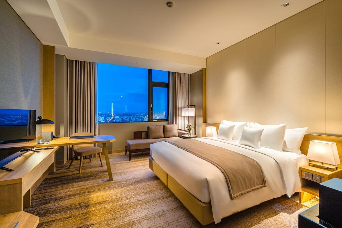 Ein Hotelzimmer mit einem großen Bett, einem Schreibtisch, einem Sessel, einem Fernseher und Blick auf die Stadtlandschaft durch das Fenster.