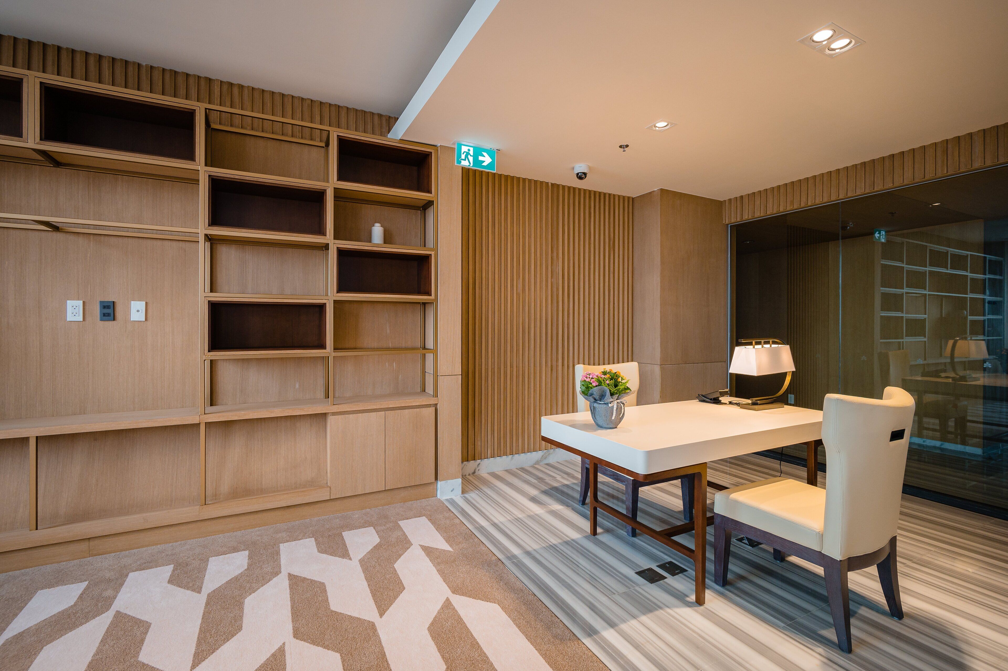 A modern office space with a wooden shelving unit, a white table with a lamp, and a patterned carpet.