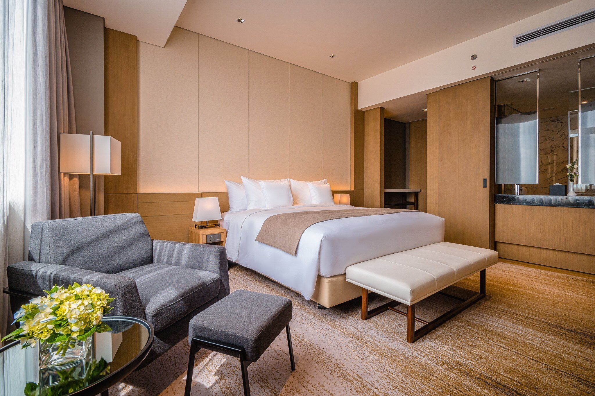 Une chambre d’hôtel moderne avec un grand lit, un canapé, une petite table ornée d’un vase de fleurs et un grand miroir.