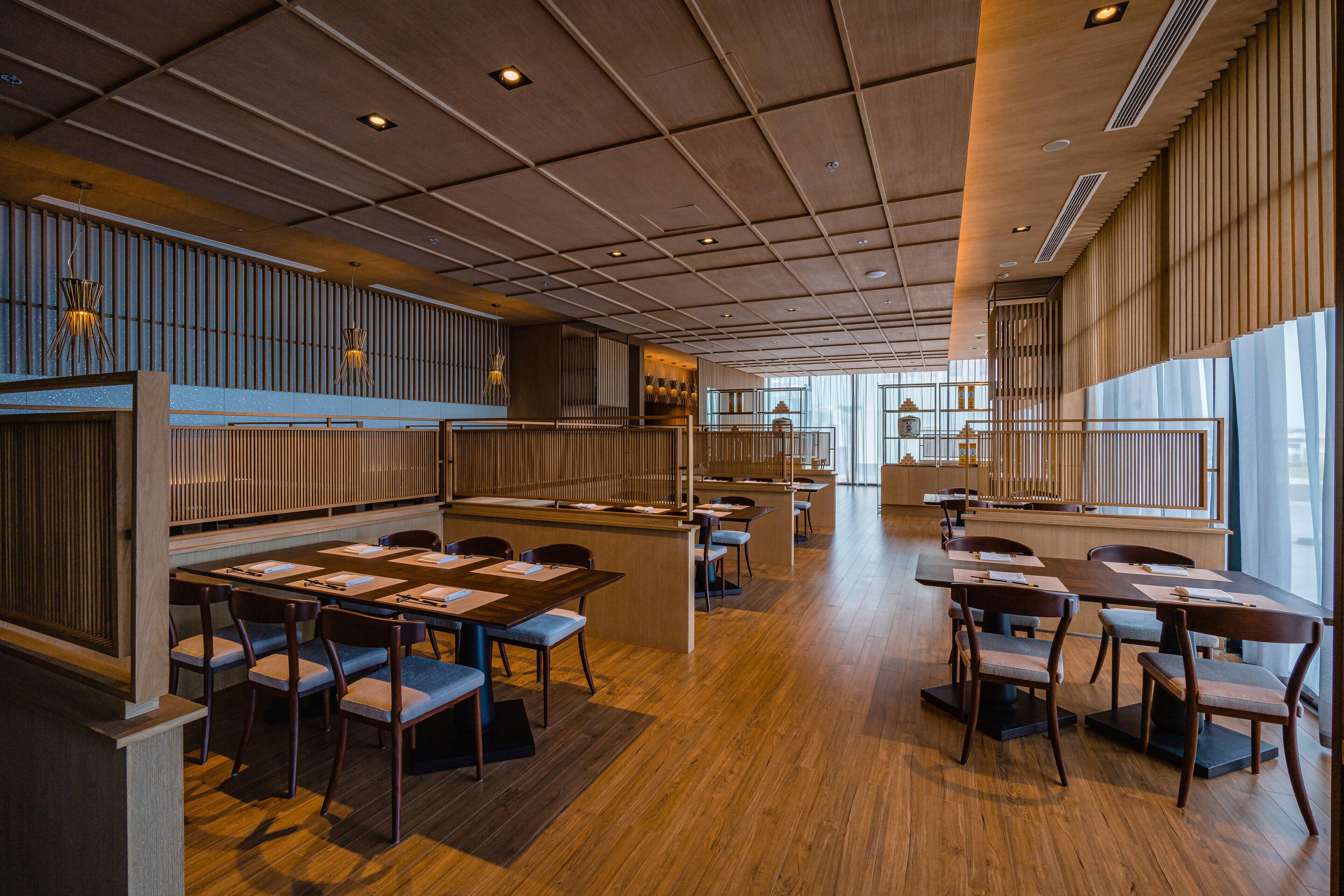 A spacious restaurant with wooden tables and chairs, a central bar area, and large windows allowing natural light.