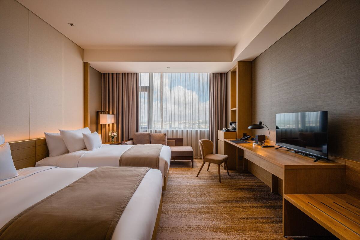 Ein Hotelzimmer mit zwei Betten, einem Schreibtisch mit Fernseher, einem Sessel und einem großen Fenster mit Vorhängen.