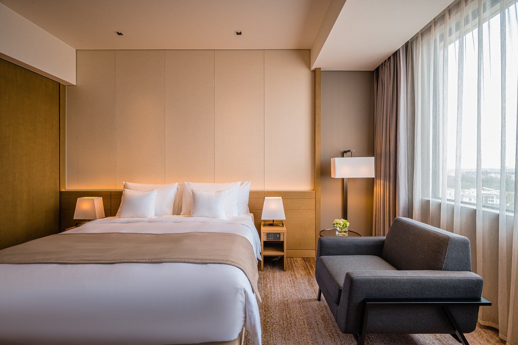 A hotel room with a bed, bedside table with lamp, chair, and large window with curtains.