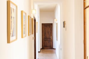 A hallway with framed pictures on the walls, a wooden door at the end, and wall-mounted lights.