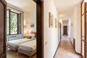 Une chambre d’hôtel avec deux lits, une fenêtre avec des rideaux, un radiateur et un couloir orné de tableaux.