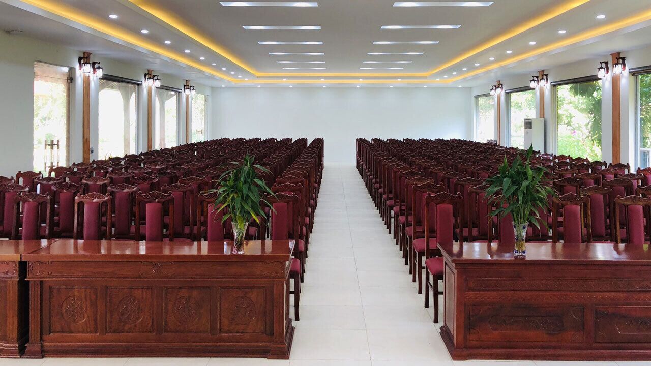 A conference room with rows of wooden chairs and tables, large windows, and ceiling lights.