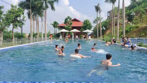 A swimming pool with people enjoying the water, surrounded by palm trees and a building with a red roof in the background. - Yasmin Farm (Linh Son)