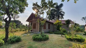 A stone house with a sloped roof, surrounded by a well-maintained garden with various shrubs and a palm tree. - Yasmin Farm (Linh Son)