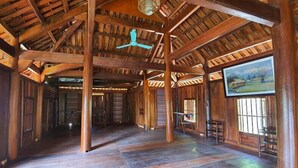 A spacious room with wooden beams, a ceiling fan, and a large window with a painting. - Yasmin Farm (Linh Son)