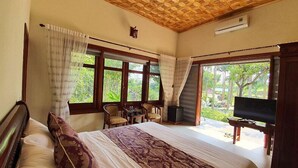 A bedroom with a large bed, a TV, a small table, and chairs. There are windows with curtains and a view of greenery outside. - Yasmin Farm (Linh Son)