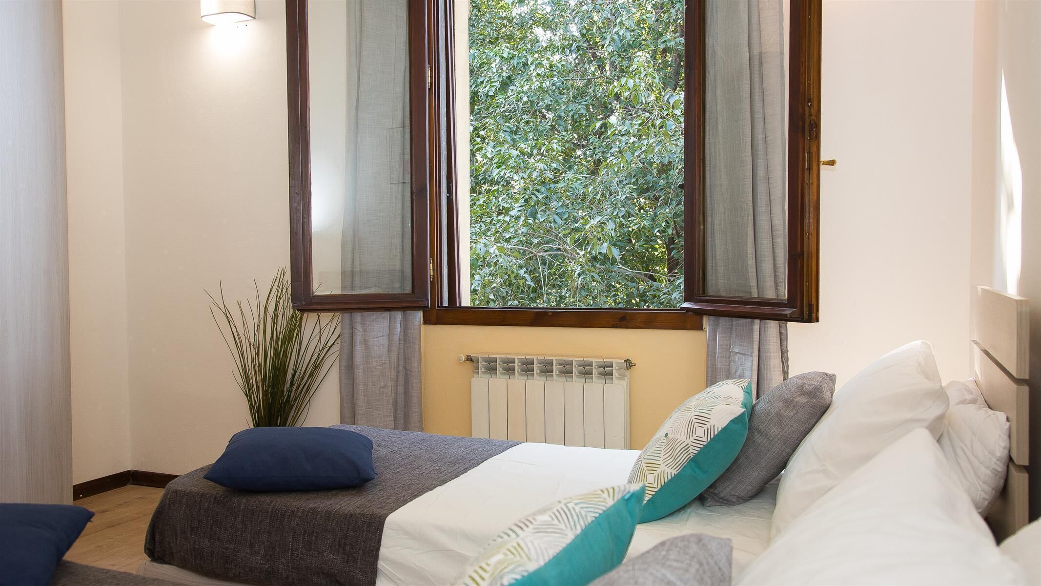 A bedroom with a bed, a radiator, a window with curtains, and a potted plant.