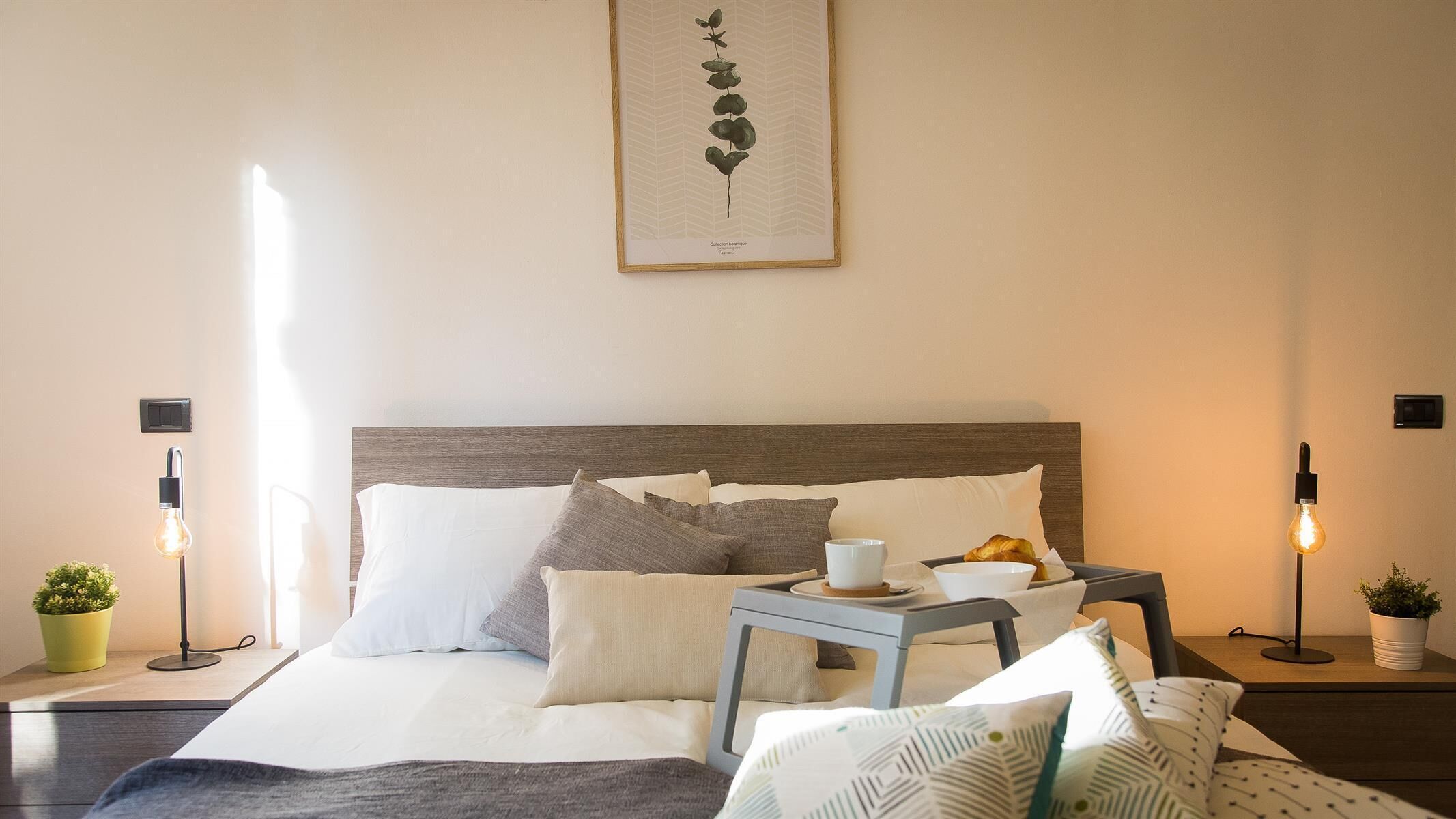 A neatly arranged bedroom with a bed, pillows, a bedside table with a cup and croissant, and a wall-mounted light fixture.