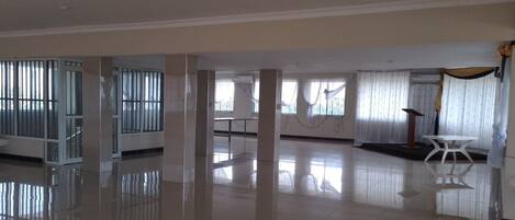 A spacious, well-lit room with a polished floor, large windows with blinds, and a white table with a chair.