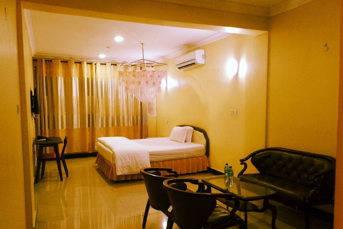 Una habitación de hotel con cama, una mesa, dos sillas, un banco, un equipo de aire acondicionado y una araña.
