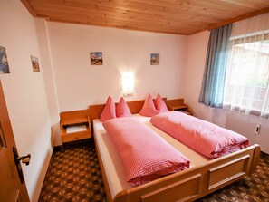 Una cama doble con ropa de cama a cuadros rojos, un cabecero de madera, una mesita de noche, una pequeña mesa y una ventana con cortinas azules.