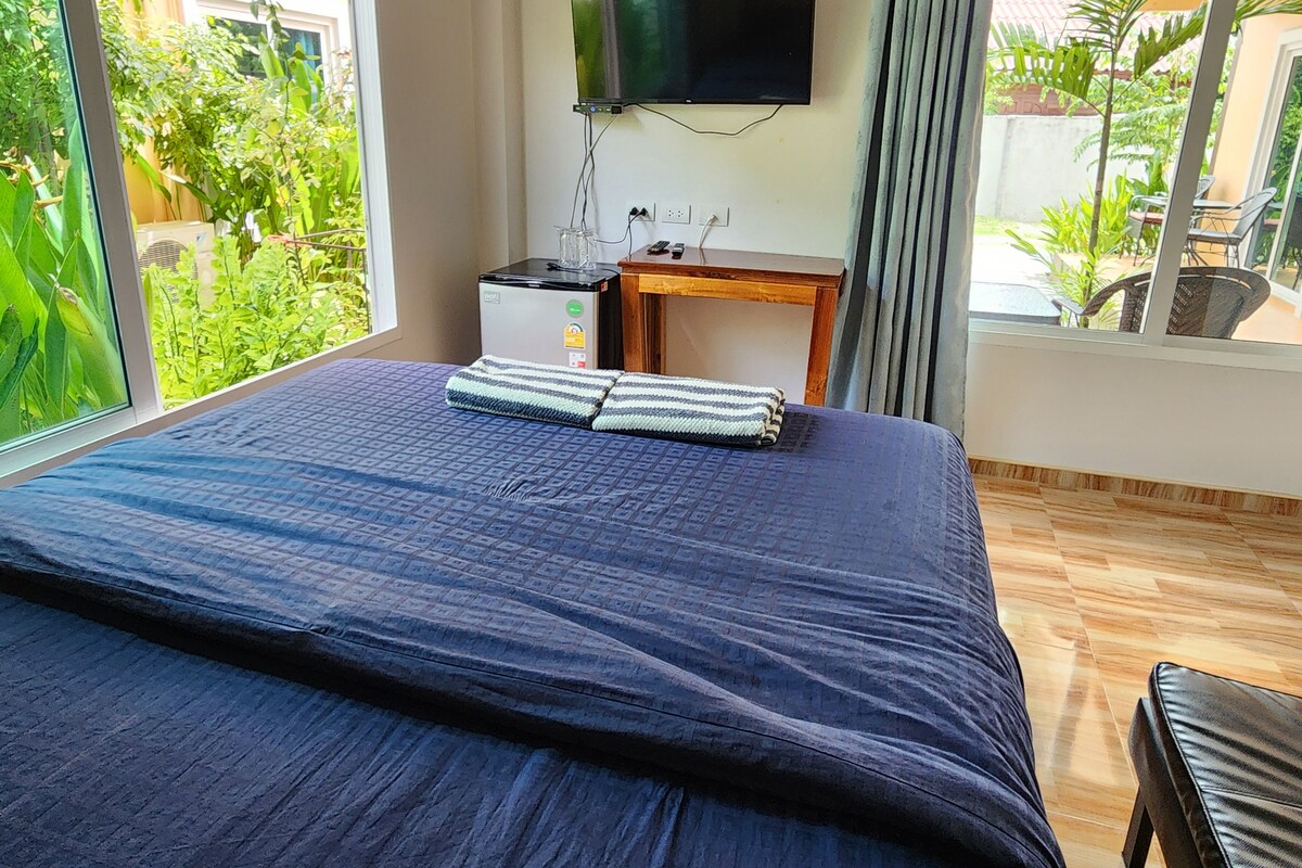 A hotel room with a bed, a desk, a television, and a view of greenery outside.