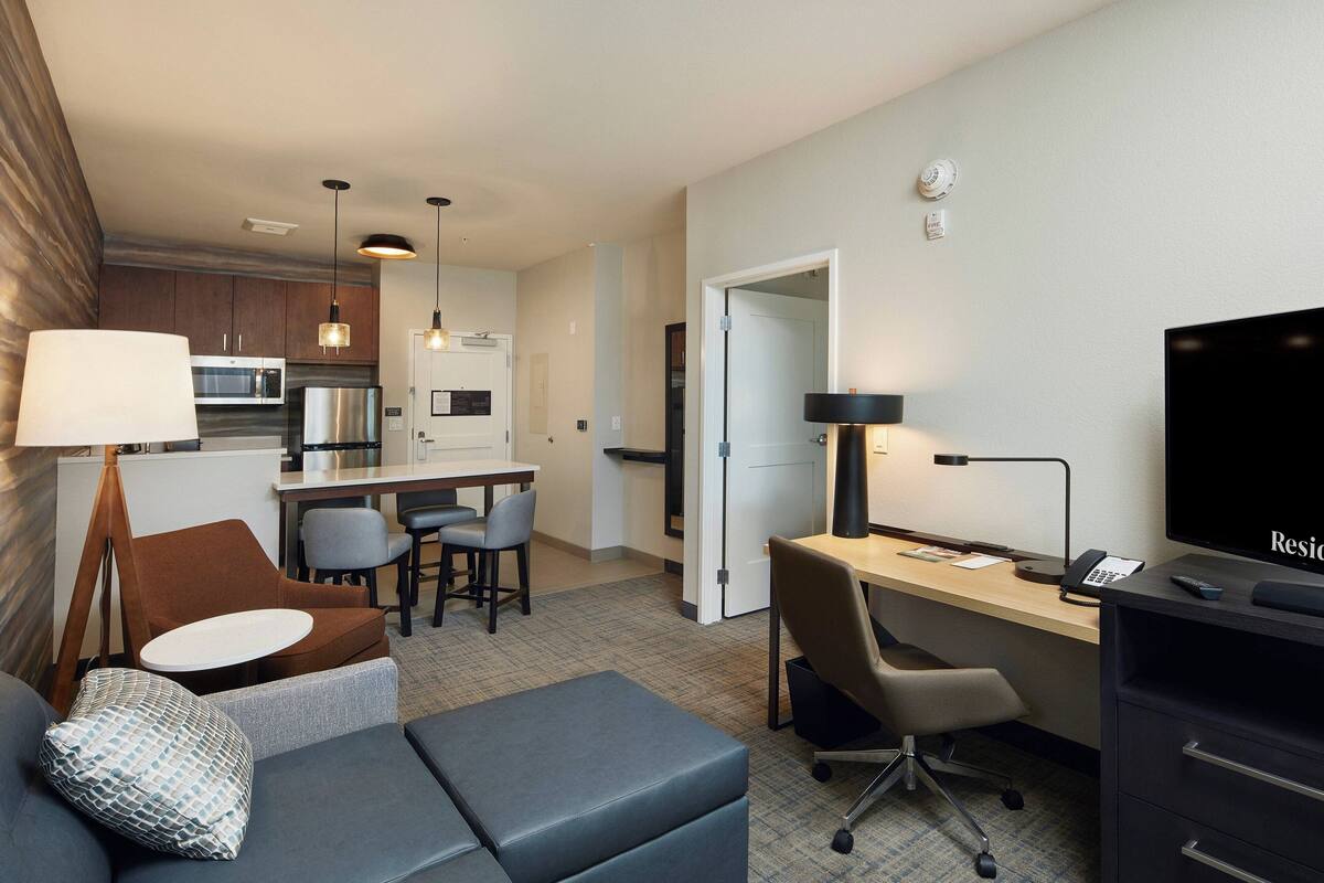 A modern hotel room with a kitchenette, a dining area, a living area with a sofa and ottoman, a desk with a chair, and a flat-screen TV.