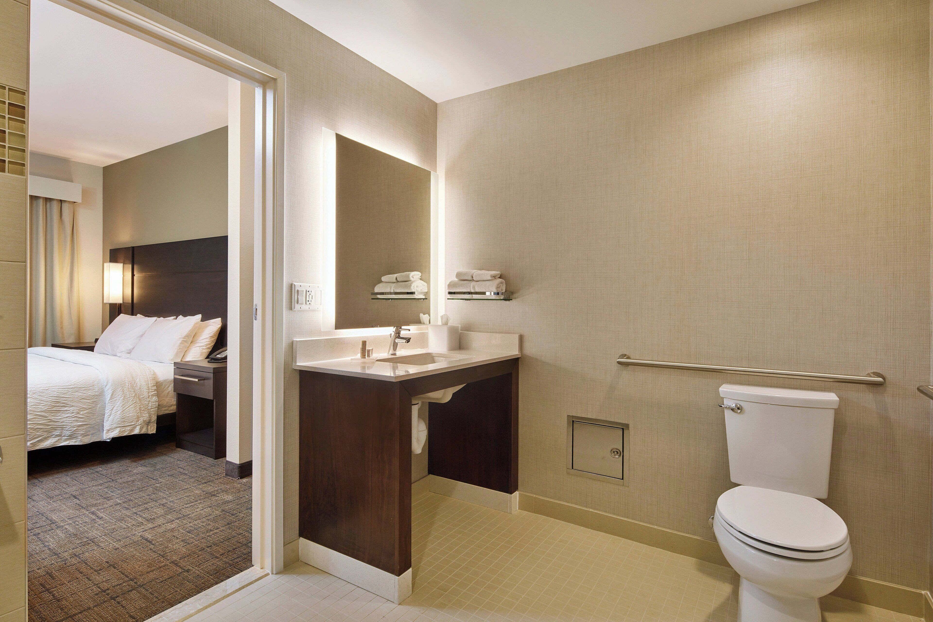 A hotel bathroom with a toilet, sink, and mirror. There is a bed in the adjacent room.