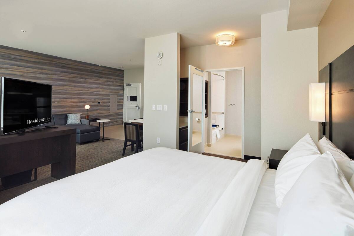 A hotel room with a large bed, a desk with a flat-screen TV, a sofa, a mirror, and a bathroom with a sink and toilet.