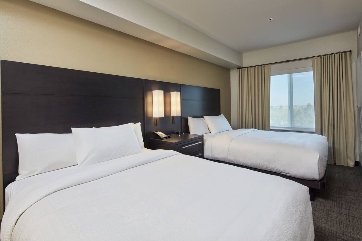A hotel room with two beds, a dark headboard, bedside lamps, a nightstand, and a large window with curtains.
