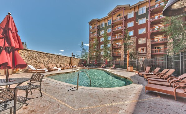 A pool area with lounge chairs and umbrellas, a stone wall, and a multi-story building with balconies.