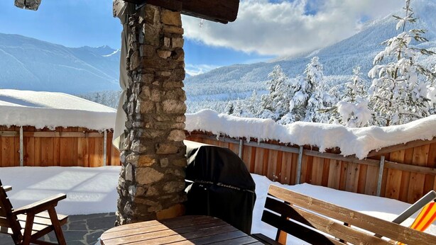 Une terrasse extérieure en bois, avec un pilier en pierre, un barbecue et une table sur laquelle se trouve une tasse.