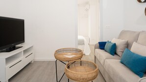 Een moderne woonkamer met een flatscreen televisie op een witte standaard, twee rieten bijzettafeltjes, een beige bank met blauwe kussens, en een witte deur naar een badkamer.