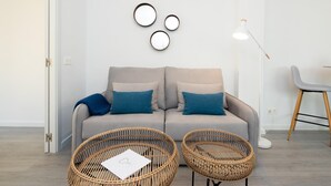 Een moderne woonkamer met een grijze bank, twee blauwe kussens en een witte muur met drie ronde spiegels. Twee rieten fauteuils staan er met een witte tafel ervoor, en een vloerlamp aan de rechterkant.