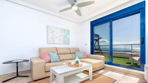 Ett modernt vardagsrum med en beige soffa, ett vitt soffbord och utsikt över havet genom ett stort fönster med blå ram.