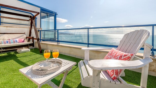 En balkong med utsikt över havet, en solstol, ett bord med ett bricka och glas, och en bänk med kuddar.