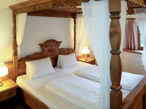 Una cama con dosel con ropa de cama blanca y cabecera de madera, dos mesitas de noche con lámparas, y una ventana con cortinas.