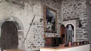 Une pièce aux murs de pierre, avec une porte en bois, une épée appuyée contre le mur, un tableau encadré représentant un cerf et un buffet en bois contenant des casseroles en cuivre.