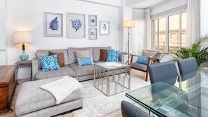 Een moderne woonkamer met een grijze bank, gestreepte kussens, een glazen salontafel en een eethoek met een glazen tafel en stoelen.
