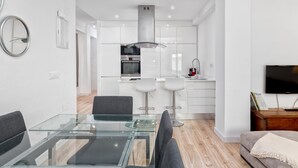 Een modern appartement met een glazen eettafel, grijze banken, een televisie en een keuken met witte kasten.