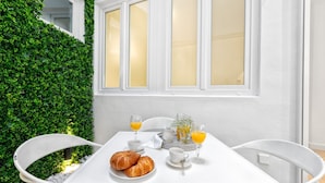 Una mesa blanca con cruasanes, tazas de café y vasos de jugo de naranja, rodeada de sillas blancas, frente a una pared blanca y un seto verde.