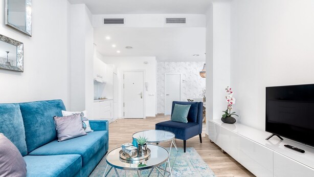 Een moderne woonkamer met een blauwe bank, een ronde glazen salontafel, een blauwe fauteuil en een flatscreen televisie op een witte kast.