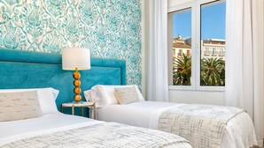 Hotelkamer met twee bedden, een muur met een blauwgroen patroon, een raam met uitzicht op gebouwen en palmbomen, en een kleine tafel met een lamp.