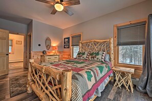 A rustic bedroom with a wooden bed frame, a quilt, a nightstand, a mirror, and a window with a view of trees. - Rustic 3-Story Pittsburg Cabin w/ Lake & Mtn Views (Pittsburg)