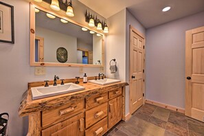 A bathroom with wooden cabinetry, two sinks, a large mirror with lights, and a door. - Rustic 3-Story Pittsburg Cabin w/ Lake & Mtn Views (Pittsburg)