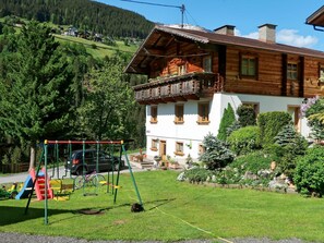 Un chalet en bois doté d’un balcon, entouré d’une végétation luxuriante et d’une aire de jeux.