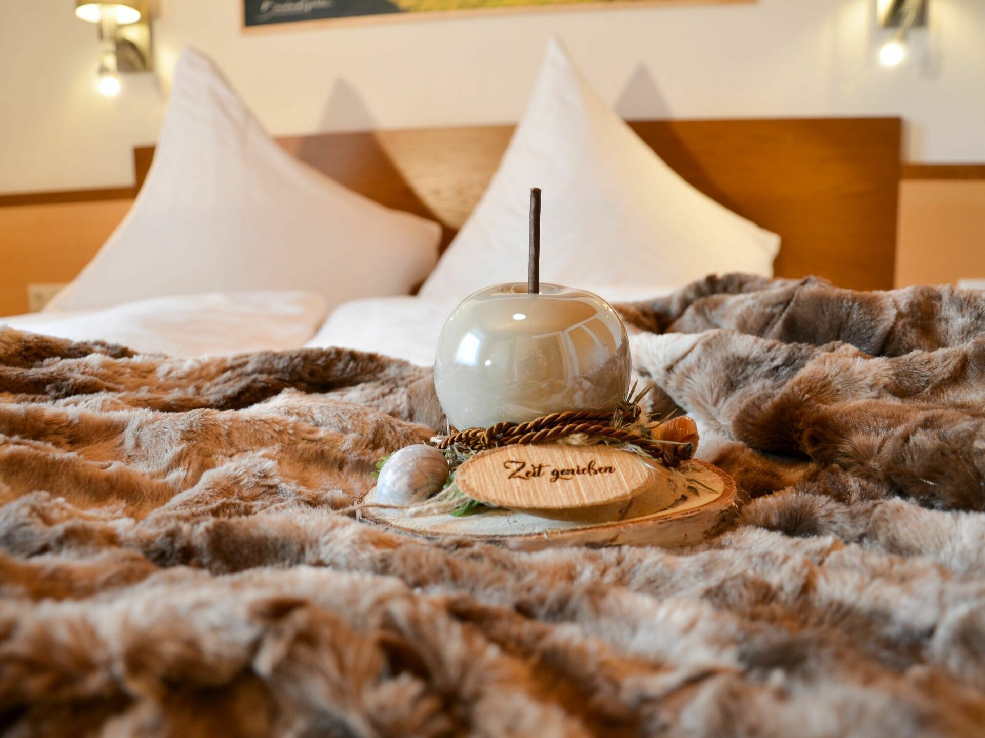 A bed with a fluffy blanket, a wooden coaster with the text "Zeit genießen," and a clear glass candle holder.
