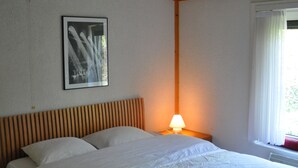 A bedroom with a wooden headboard, a single bed, a nightstand with a lamp, a framed picture on the wall, and a window with curtains. - The Happy House (De Haan)