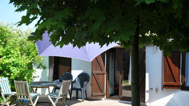 An outdoor seating area with a table and chairs under a large umbrella, a wooden door, and a white building with a window. - The Happy House (De Haan)