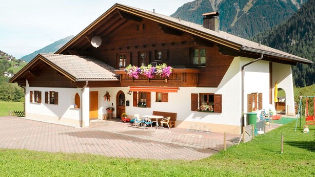 A traditional chalet with a wooden facade, a balcony with hanging flower baskets, and a paved patio area with outdoor seating. - Nice apartment for 5 guests with WIFI, TV, patio and pets allowed (Gaschurn)