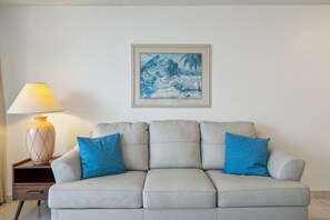 A modern living room with a light grey sofa, two blue cushions, a wooden side table with a lamp, and a framed landscape painting on the wall.