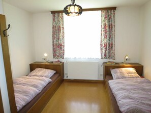 A room with two beds, a radiator, a window with floral curtains, and a ceiling light fixture.